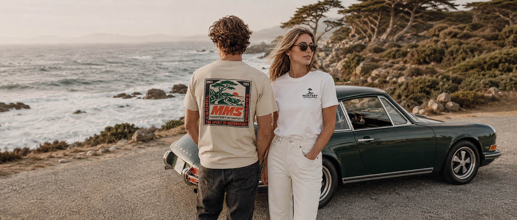 A man and a woman standing outdoors by the ocean next to a dark green classic car, wearing MMS branded t-shirts. The man's shirt features a large graphic print on the back, and the woman wears a smaller chest logo.